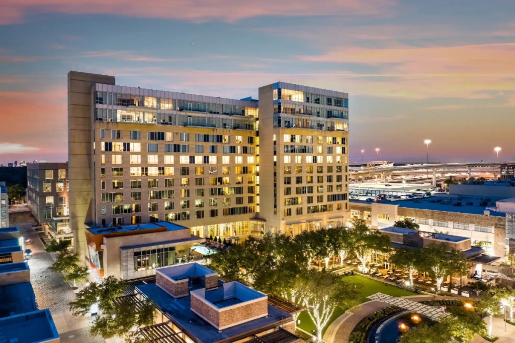 exterior of a hotel in Houston at sunset with some lights in some guest rooms and a view of a lit fountain